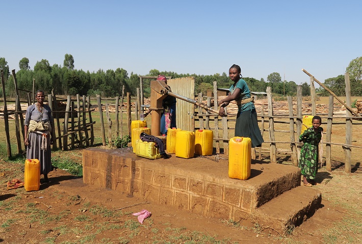 Nainen pumppaa käsikaivolla vettä keltaiseen isoon kanisteriin. Nuori tyttö kantaa selässää kanisteria. Yksi nainen seisoo kuvan reunassa.