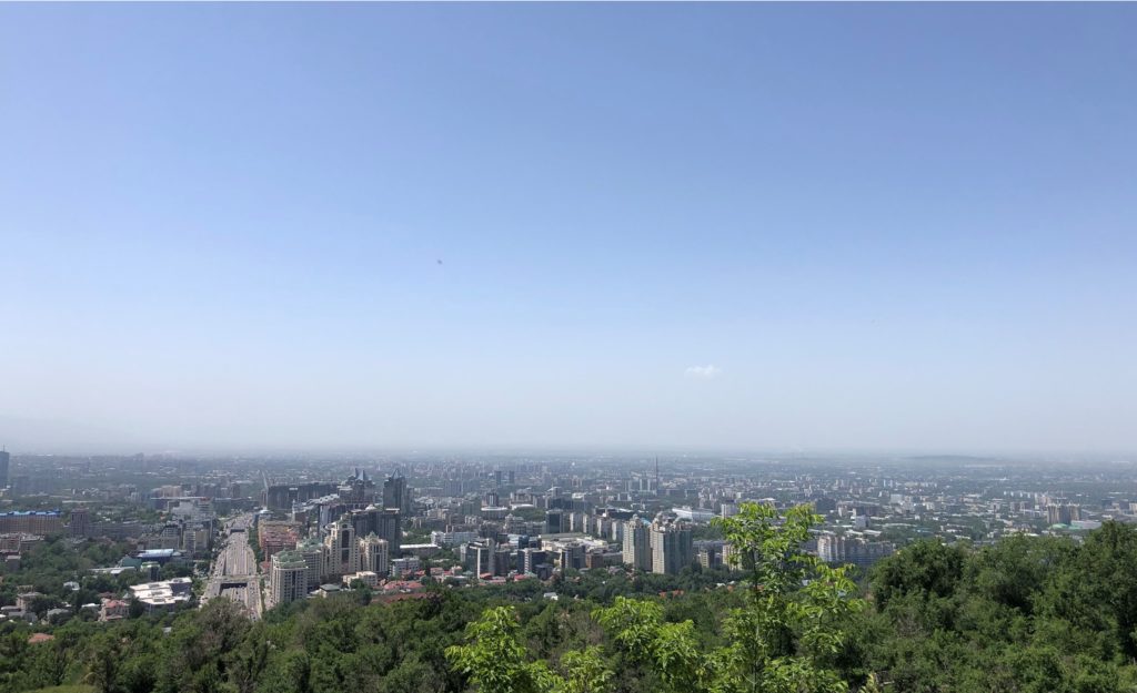 Kuvassa näkyy Almatyn kaupunki Kök Töbe-vuoren huipulta. Sää on kirkas ja aurinko paistaa.